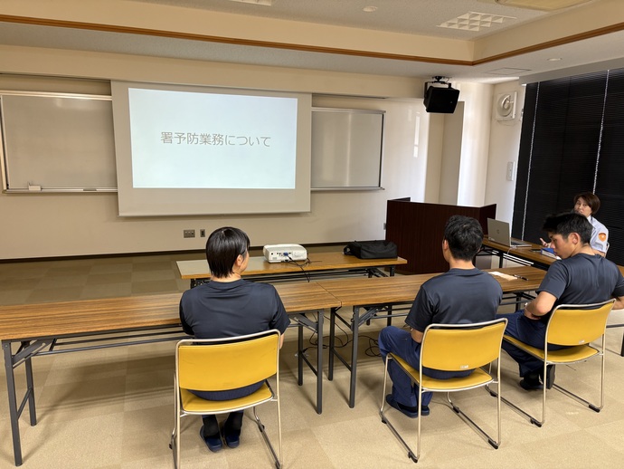 初任教育中の学生が、予防班員にプロジェクターを使用し、パワーポイント投影しながら予防業務について教えてもらっている写真。
